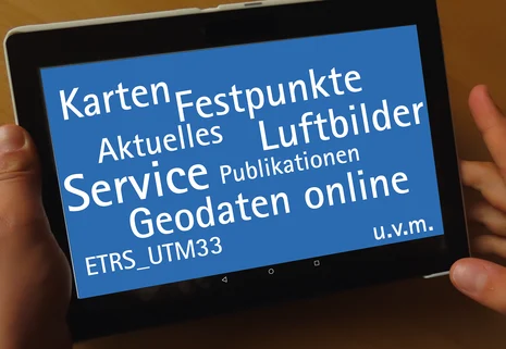 Auf dem Bild wird ein Tablet in zwei Händen gehalten. Darauf befinden sich Begriffe in weißer Schrift auf blauem Hintergrund. Sie stammen aus dem Bereich der Vermessung, wie bspw. "Karten", "Luftbilder", "Geodaten online" u.v.m..