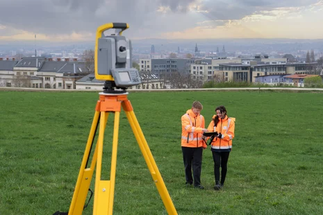 Zwei Vermesser bei der Arbeit im Hintergrund eine Stadtkulisse im Vordergrund ein Vermessungsgerät