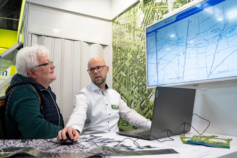Alexander Krautz vom GeoSN zeigt einer Messebesucherindas Geoportal Sachsenatlas am Laptop und auf einem großen Monitor