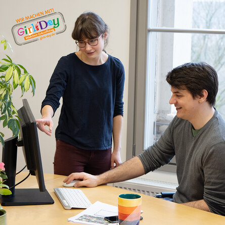 Ein junger Mann und eine junge Frau unterhalten sich über den Inhalt auf dem PC-Monotor. In der oberen rechten Ecke ist das Logo des Girls'Days 2025 zu sehen.